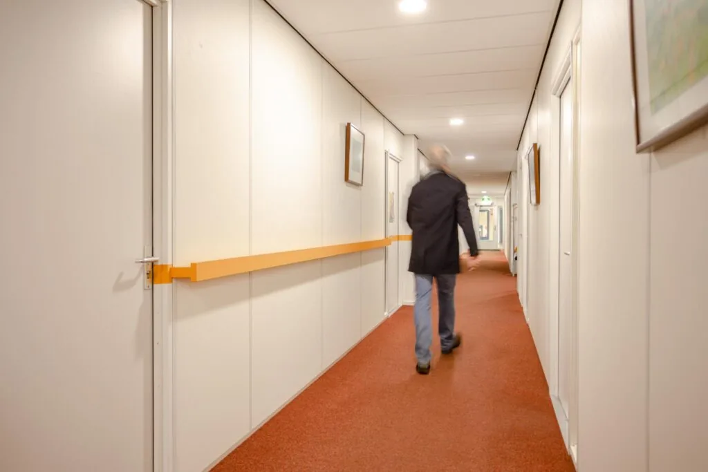 Man loopt door een gangenstelsel met witte muren, rood tapijt en oranje wandbeugels in het gebouw aan de Christiaan Krammlaan.